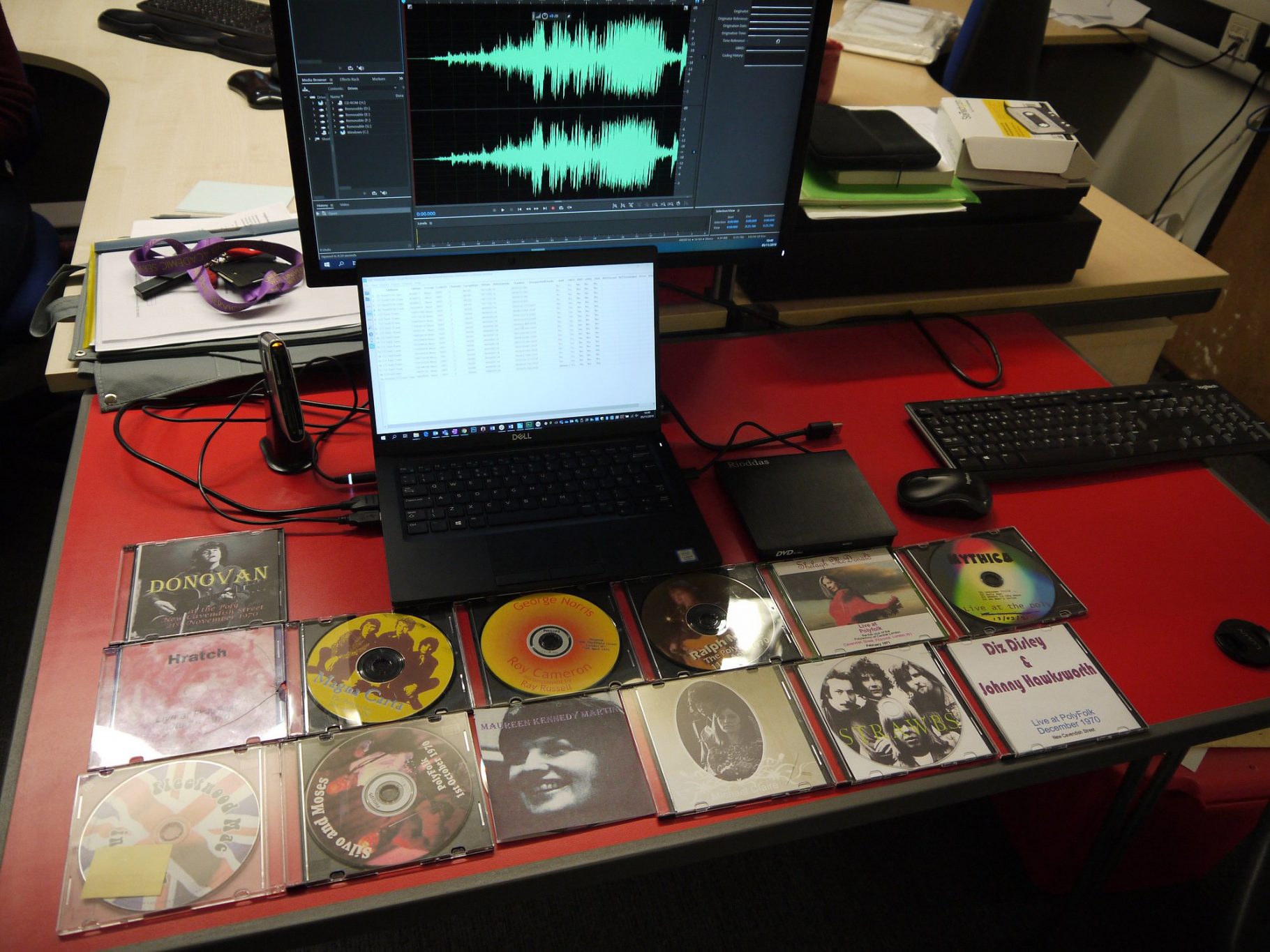 A photograph of a set of audio CDs awaiting digitisation. Behind the CDs is a computer, on the computer screen is an audio wave form.