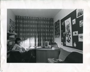 A student bedroom in the Marylebone campus. A student is seated at her desk in front of books and papers. The walls are adorned with curtains and posters..