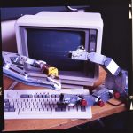 A simple robot hand holds a large die, while another looks poised to take it. In the background is a 1980s microcomputer.