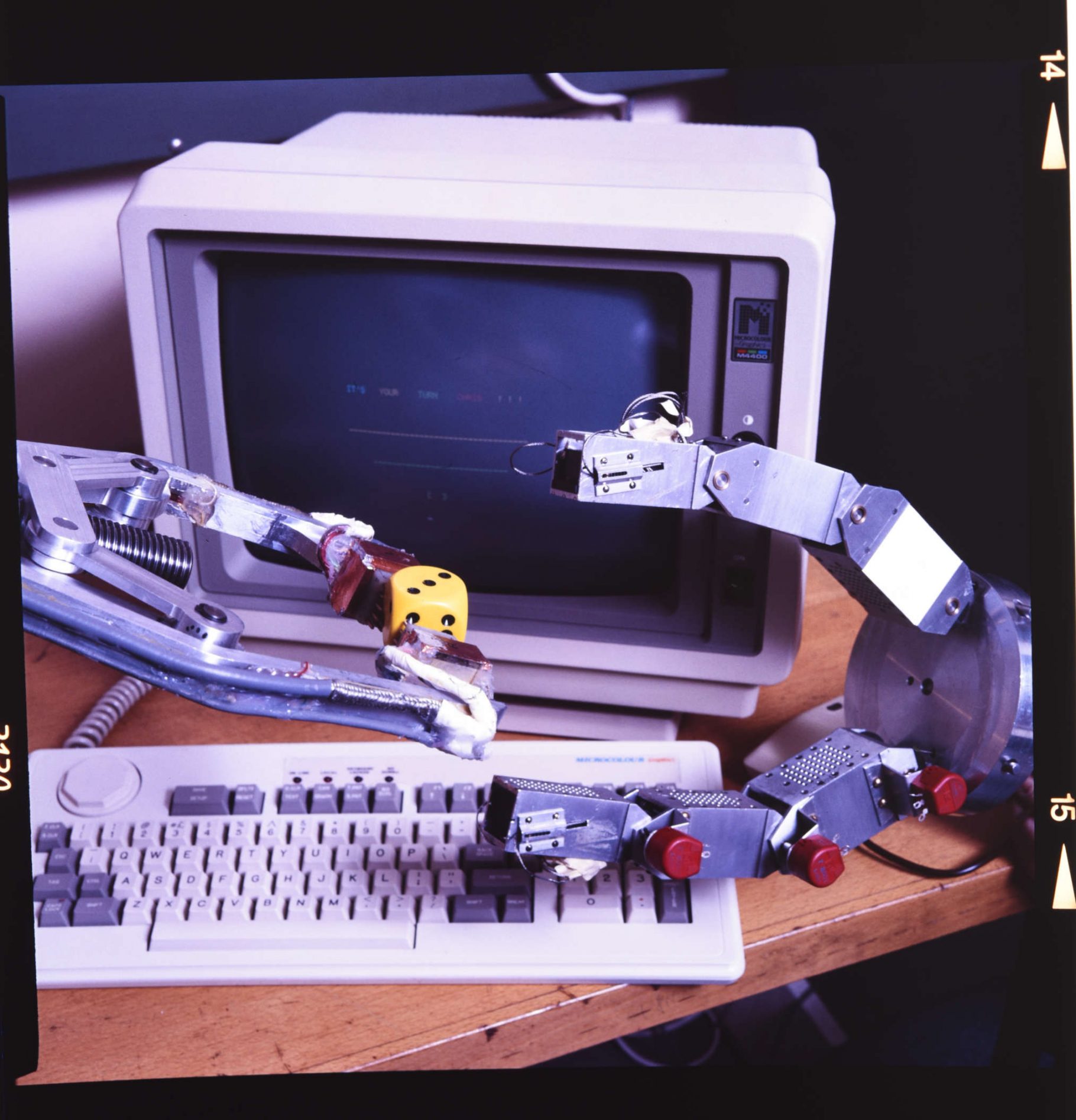 A simple robot hand holds a large die, while another looks poised to take it. In the background is a 1980s microcomputer.